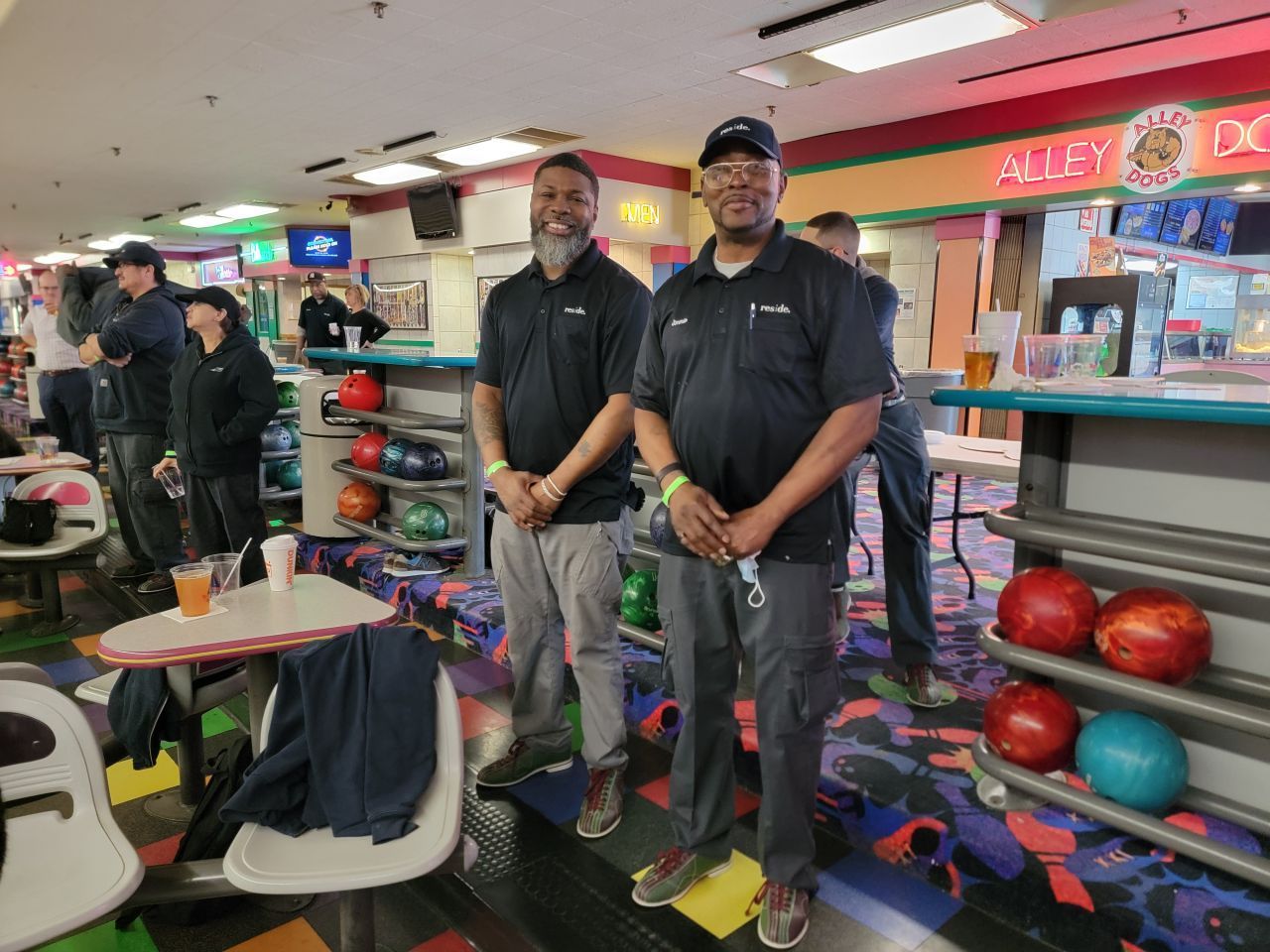 Two staff members in black shirts stand in a bowling alley beside lanes and colorful bowling balls.