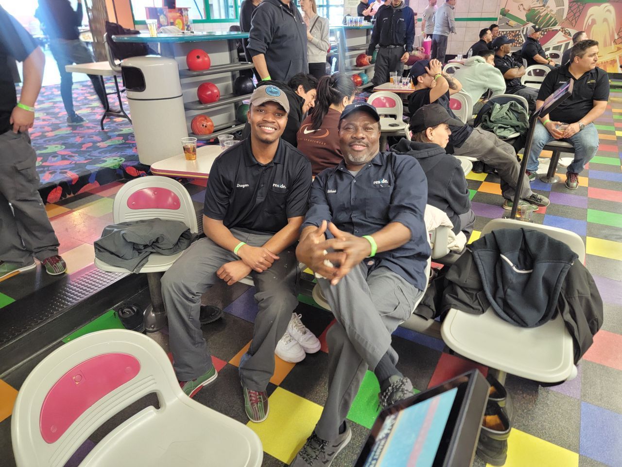 Two people sit smiling in colorful bowling alley seating with bowling shoes and people in the background