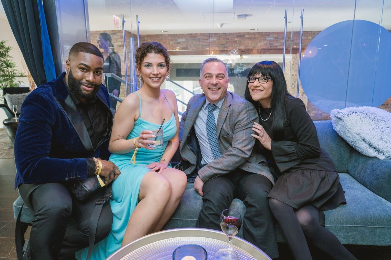 Four people seated on a couch at an event, smiling and posing with drinks in a lounge setting.