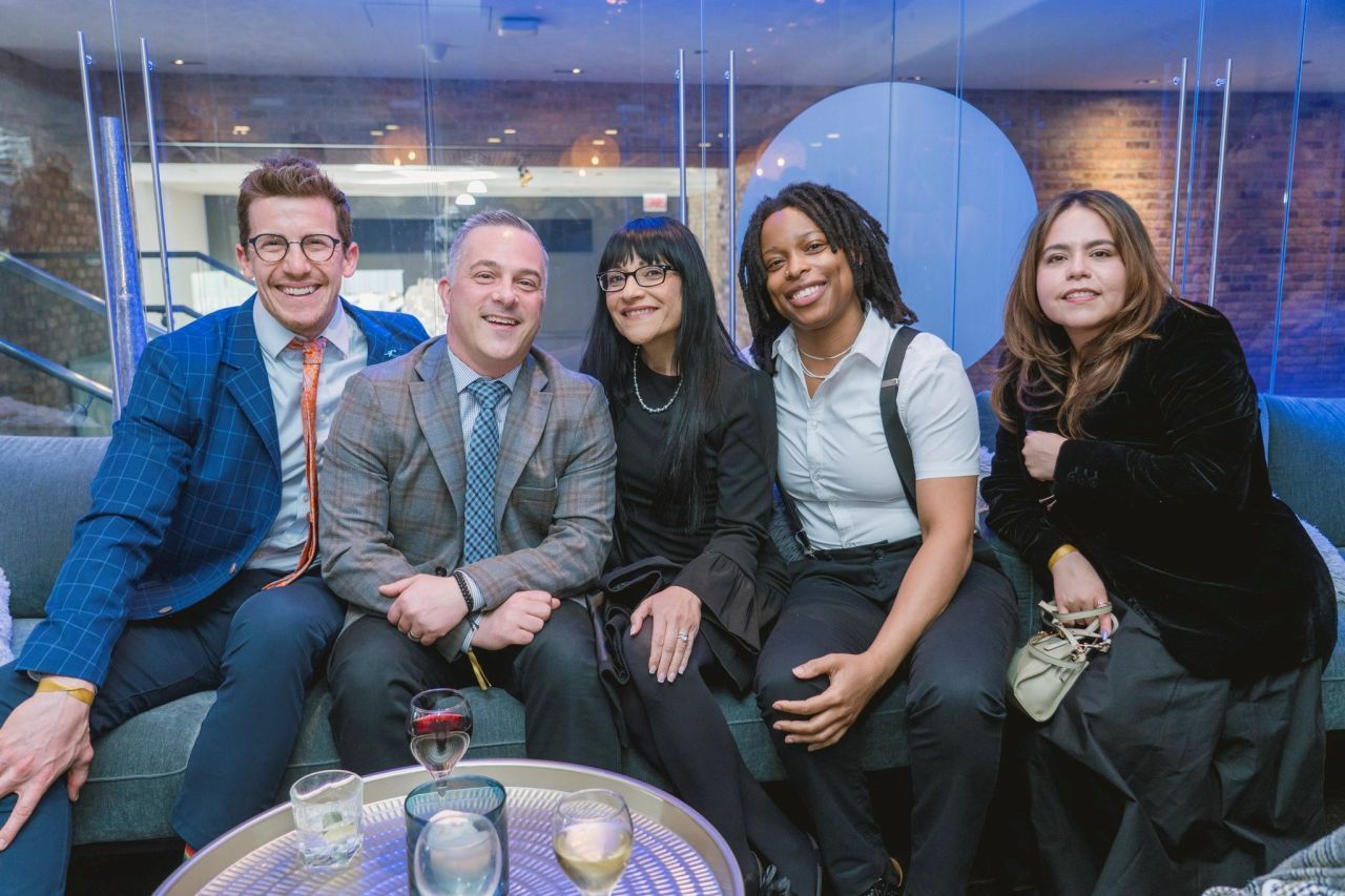 Five people smiling on a couch at a social gathering, with drinks on a table in front of them.