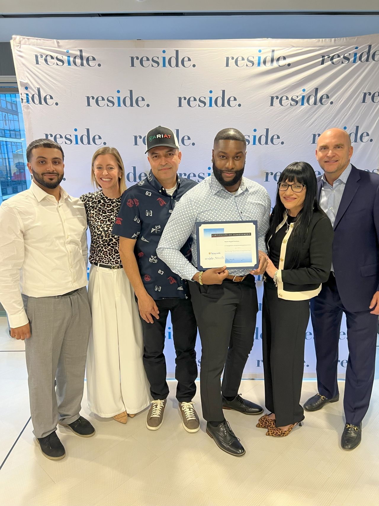 Six people posing at a reside event, one holding a certificate in front of a branded backdrop.