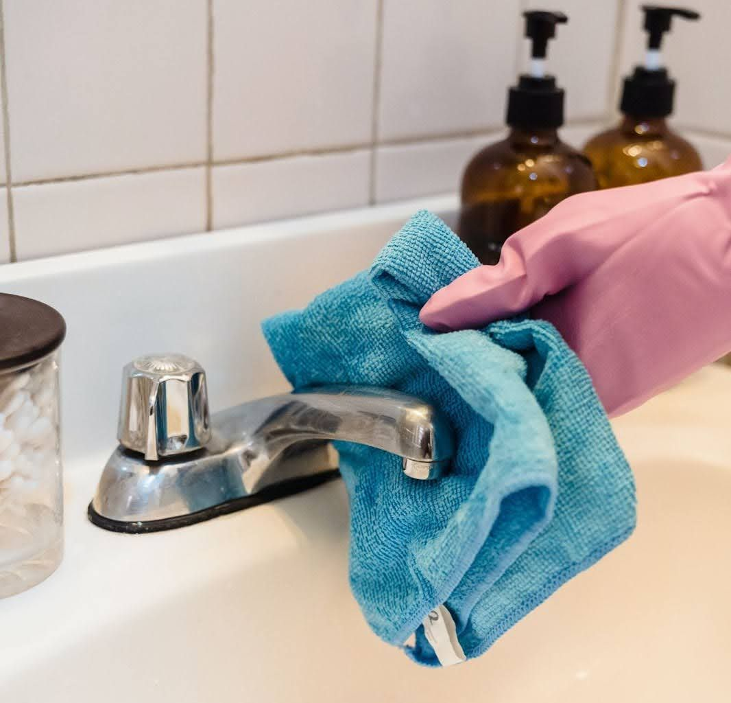 A gloved hand uses a blue cloth to wipe a silver bathroom faucet above a white sink.