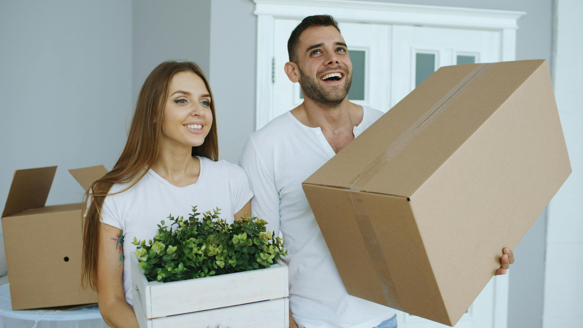 A smiling couple carrying moving boxes in a bright home, one box filled with a green plant