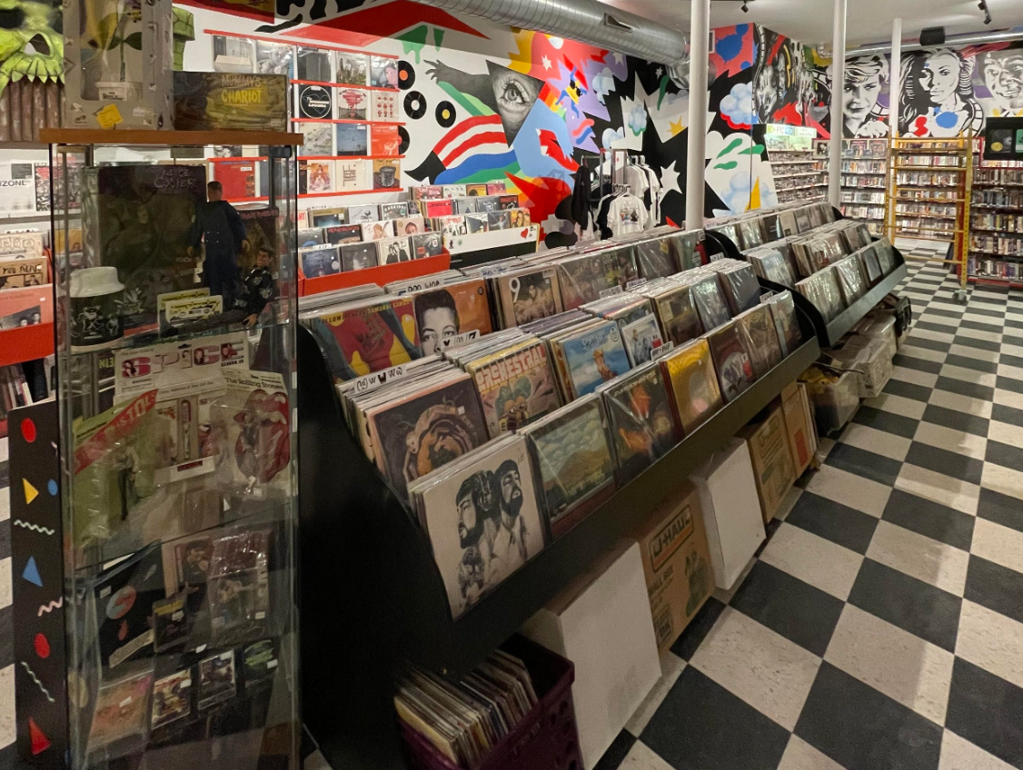 A record store with a checkered floor, rows of vinyl records in display bins, and colorful murals on the walls.