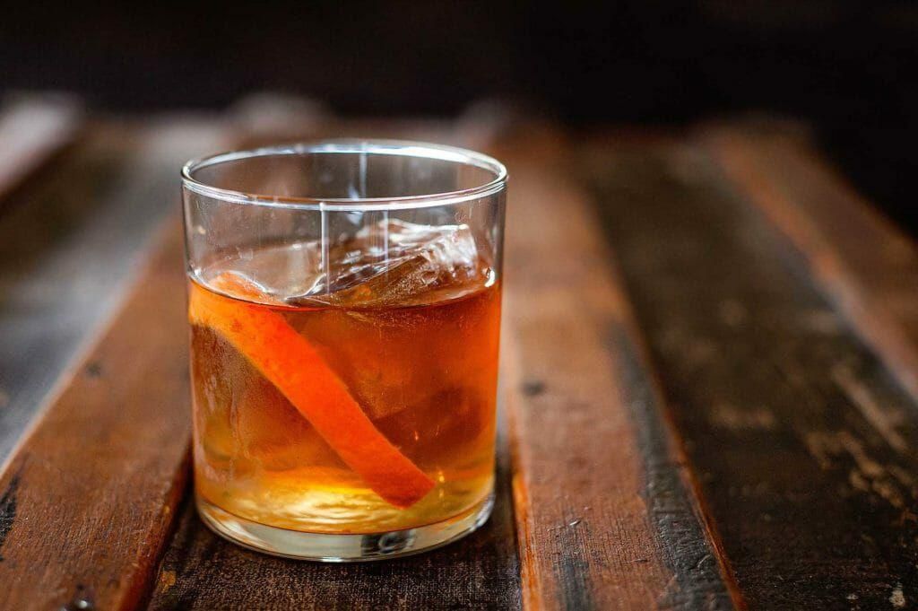 A glass of whiskey with ice and an orange peel on a wooden table.
