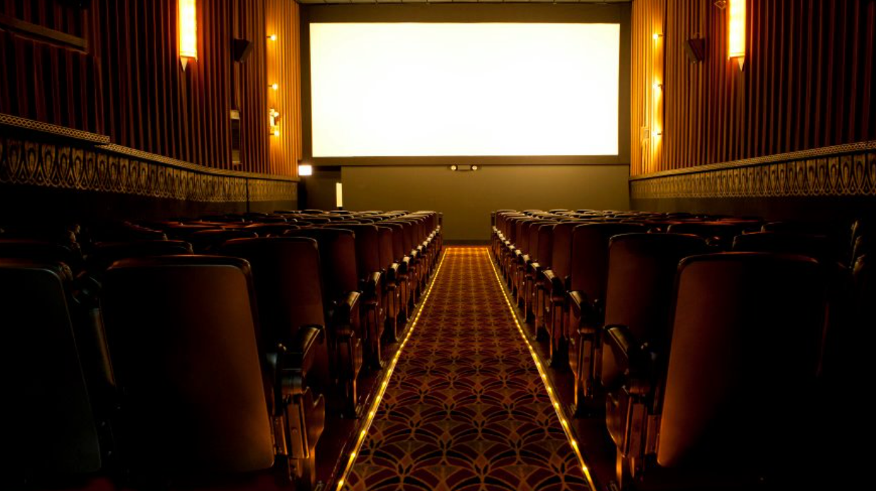 Empty movie theater with rows of seats on either side of a patterned aisle leading to a blank, illuminated screen.