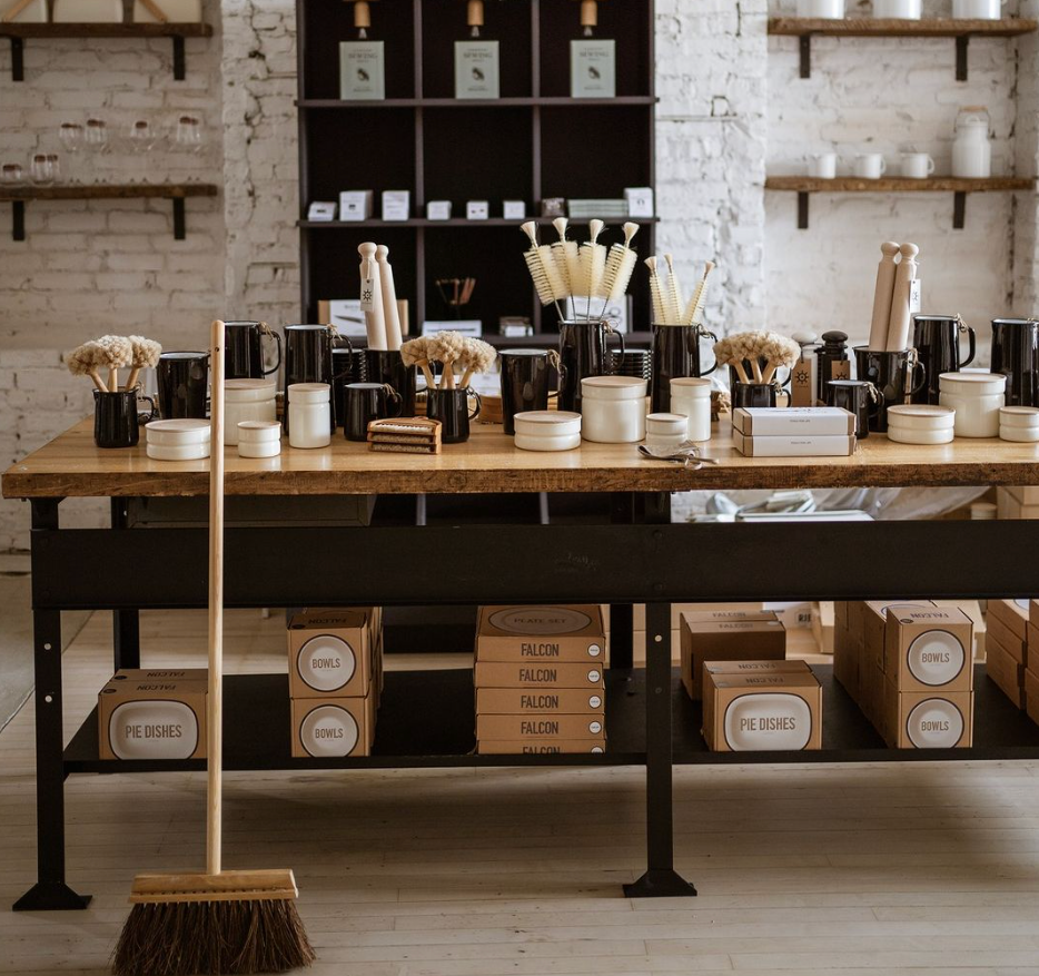 A retail store display featuring household brushes and ceramics on a wooden table, with shelves and a broom in the foreground.