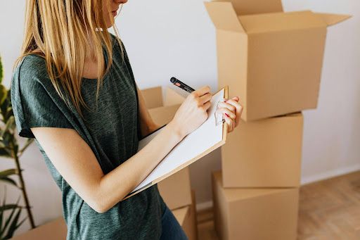 Person writing on a clipboard beside stacked cardboard moving boxes in a room