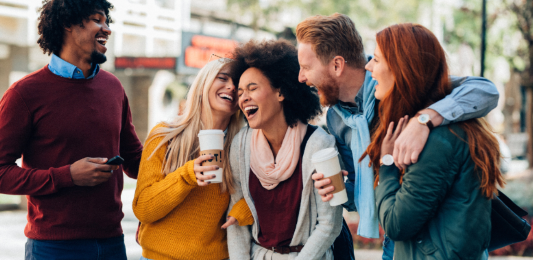 Four friends laughing together outdoors, wearing colorful sweaters and jackets, holding drinks.