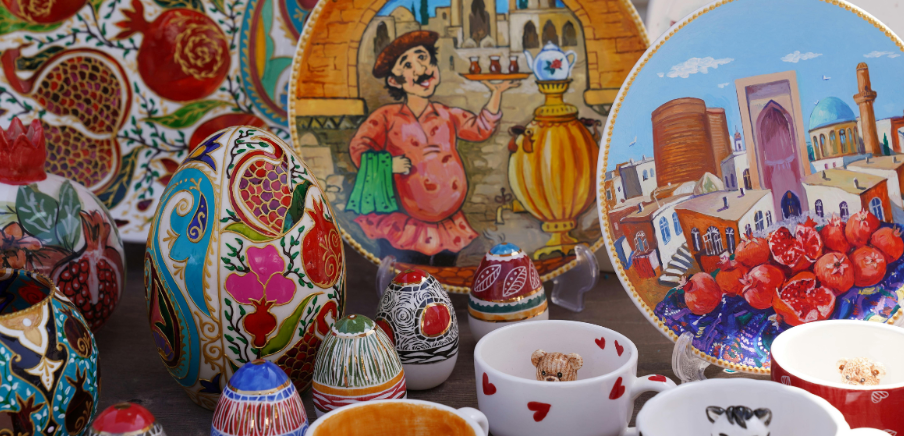 Colorful ceramics and painted souvenirs displayed with tea cups and a seaside city scene, in bright red and blue colors