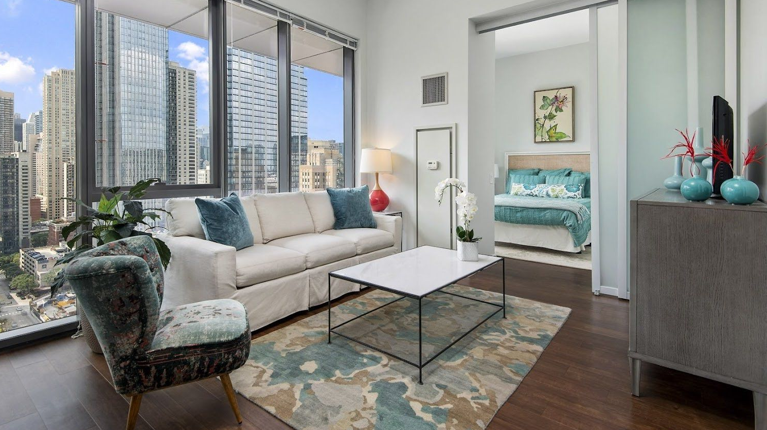 A modern living room with a white sofa, patterned armchair, coffee table, and area rug, open to a view of city buildings.