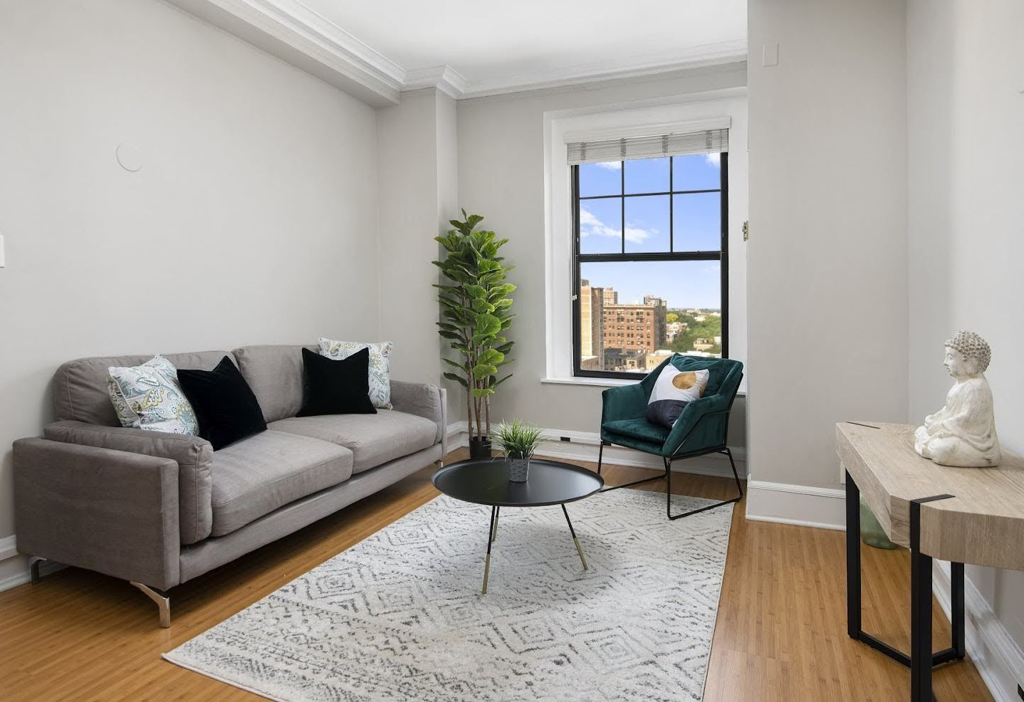 A modern living room with a grey sofa, teal armchair, round coffee table, houseplant, and white Buddha statue on a table.