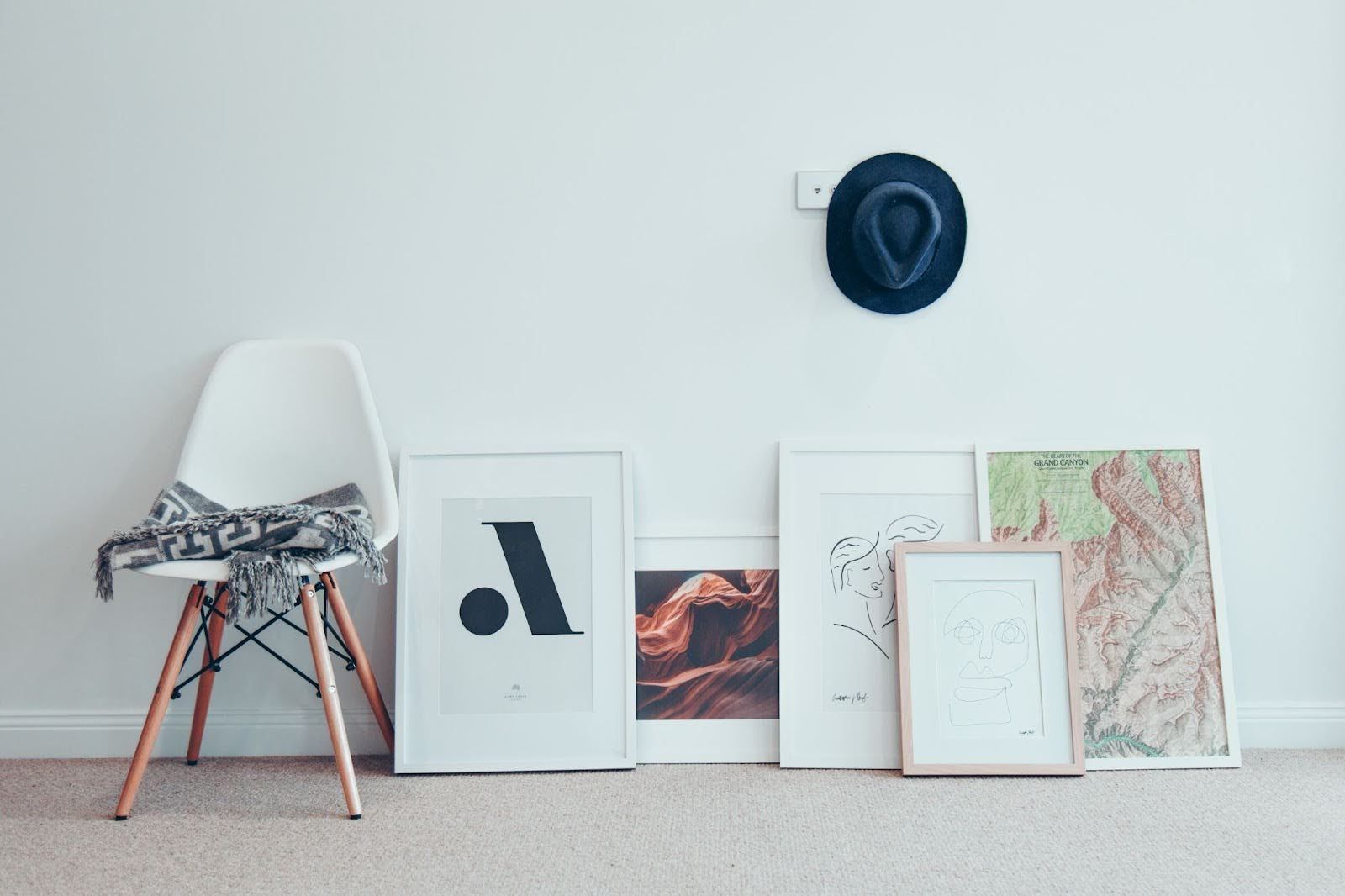 Wall with a chair and framed modern art laying agains the floor and the wall. A single fedora hat hang on a hook