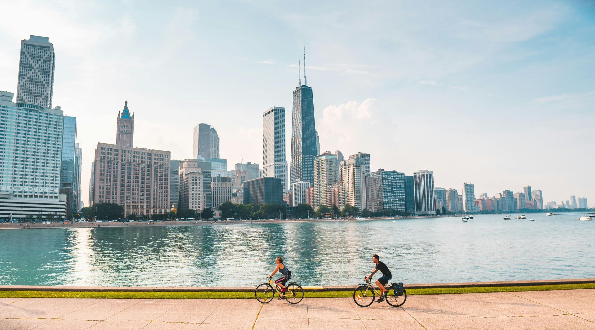 Cyclists ride along a path by water, with a city skyline of tall buildings in the background.