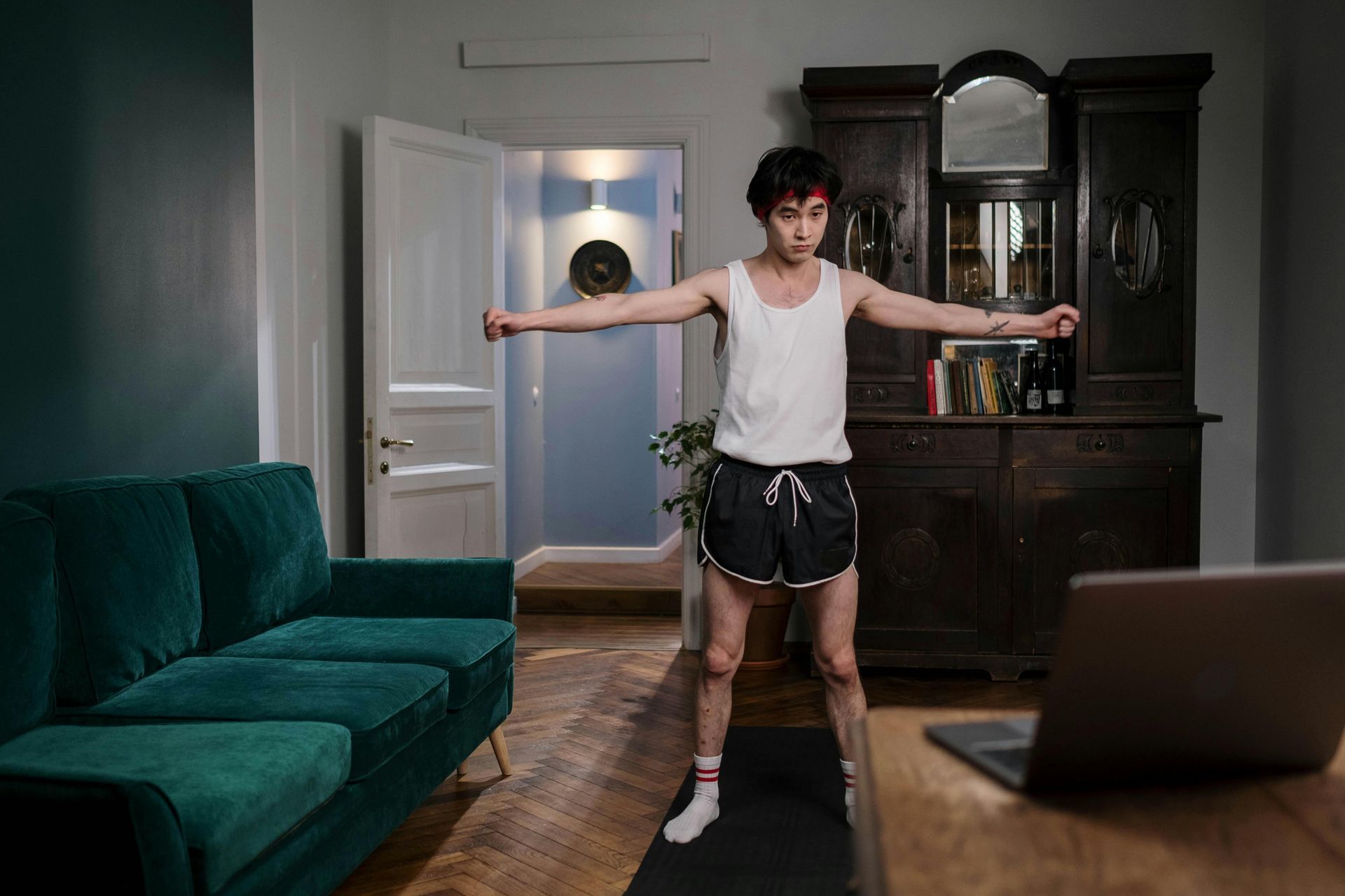 A person in a white tank top and dark shorts stands on a yoga mat in a living room, arms outstretched, following a laptop.