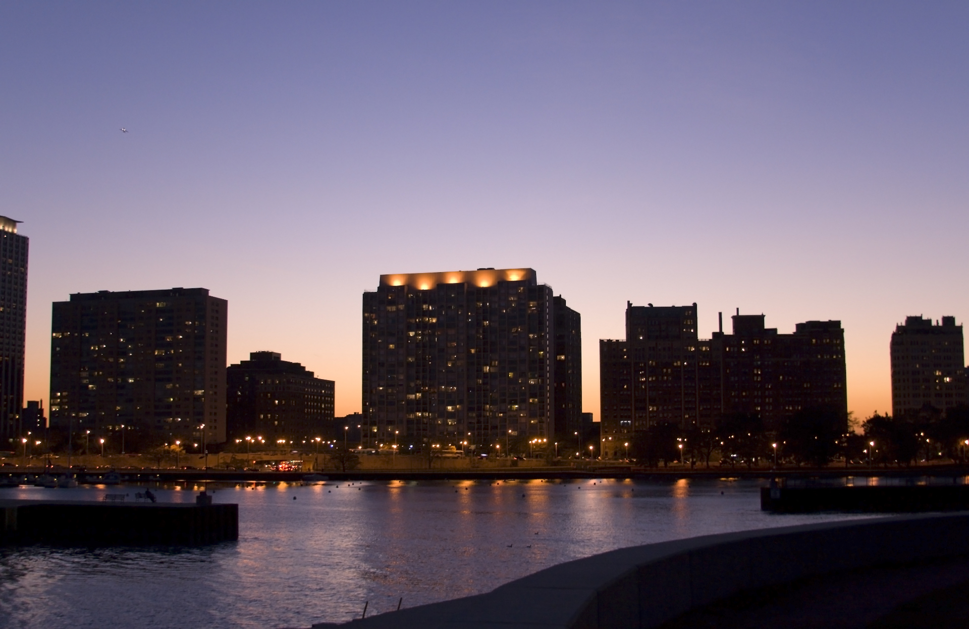 City skyline at dusk reflected in calm water, with lit buildings against a purple evening sky