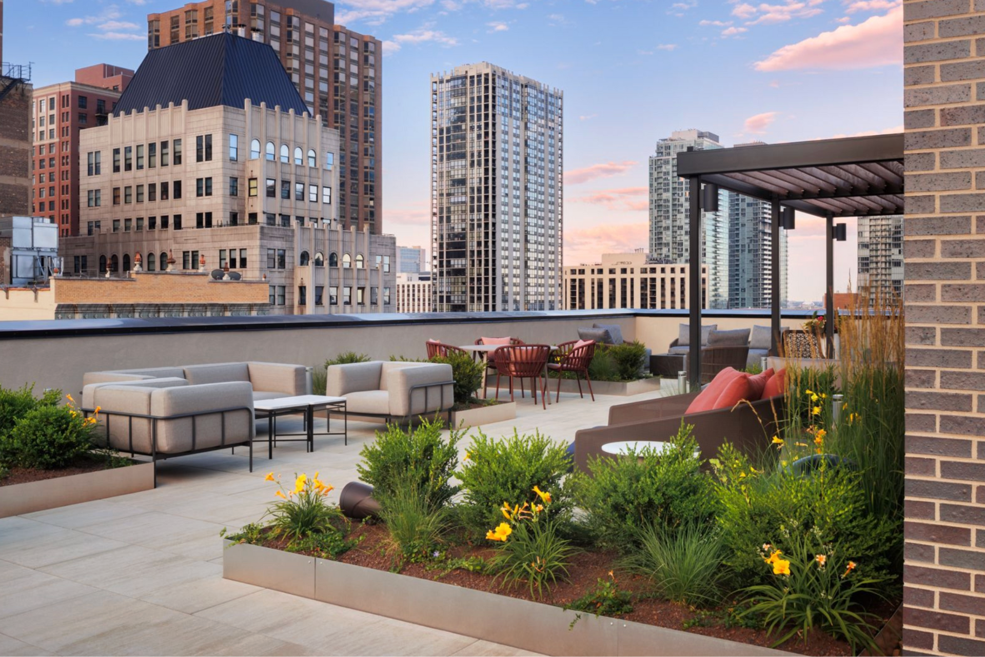 Rooftop patio with lounge seating, flower beds, and city buildings at sunset.