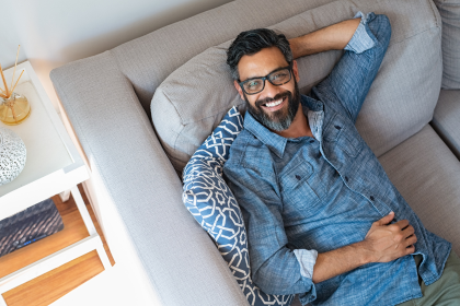 Smiling man reclining on a light gray couch with one hand behind his head, wearing glasses and a blue shirt.