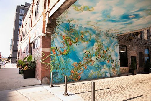 A mural depicting blue sky and green vines on the side and ceiling of an urban building, next to a cobblestone street.