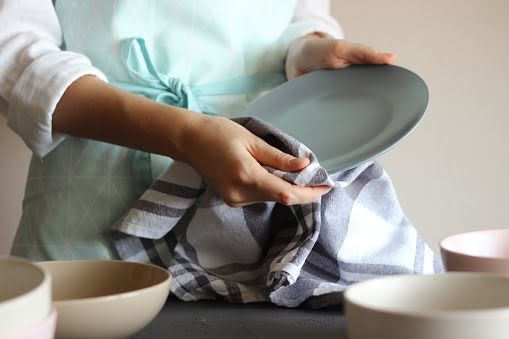 A person wearing a light blue apron uses a grey and white plaid towel to dry a grey plate, with bowls on a table.