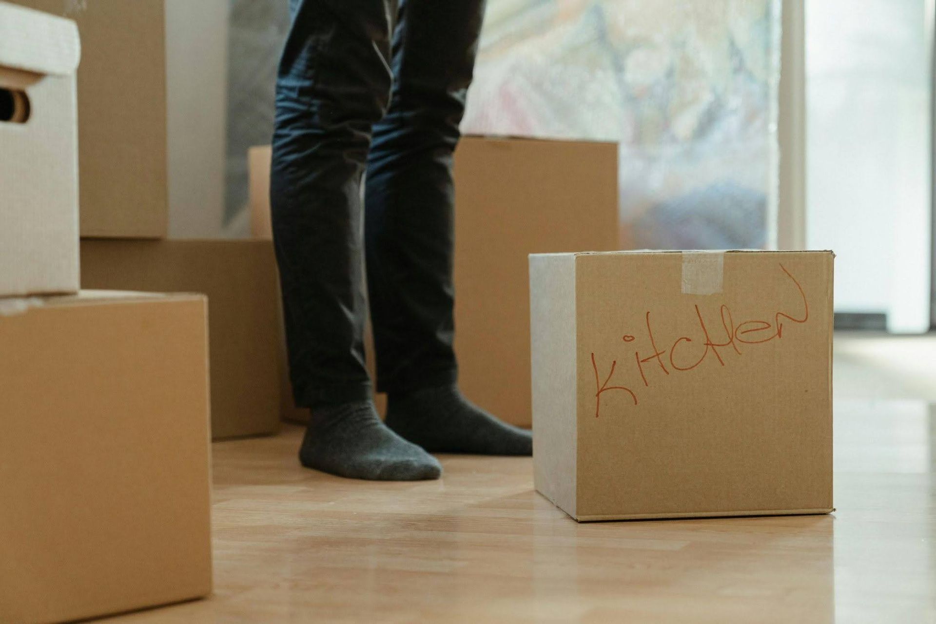 Person in black pants and socks standing among moving boxes, one labeled “Kitchen”
