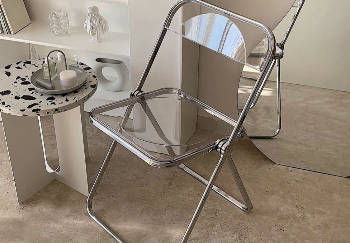 A clear acrylic folding chair with a metallic frame sits next to a small, round table with a black and white speckled top.