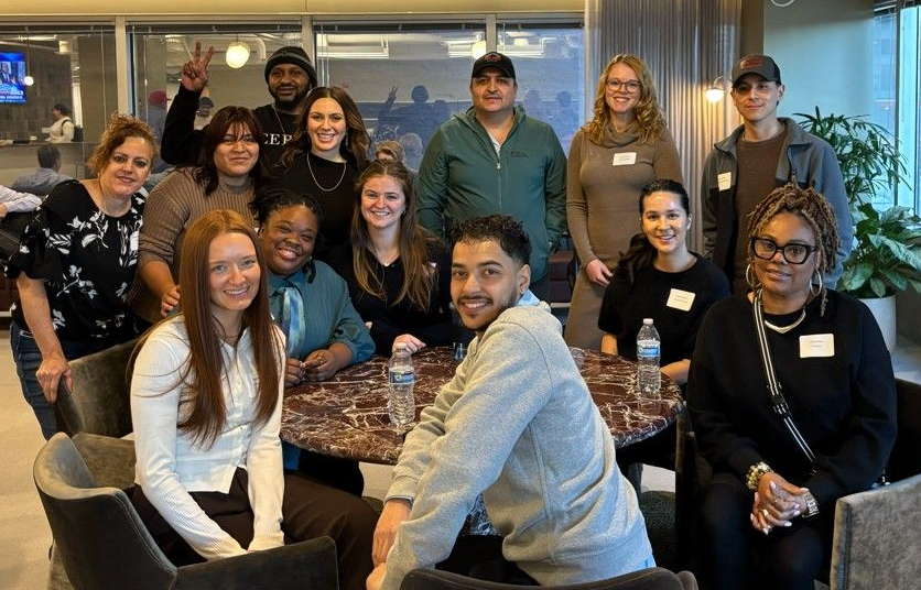 Group of people posing together in a modern office lounge, smiling around a table with chairs.