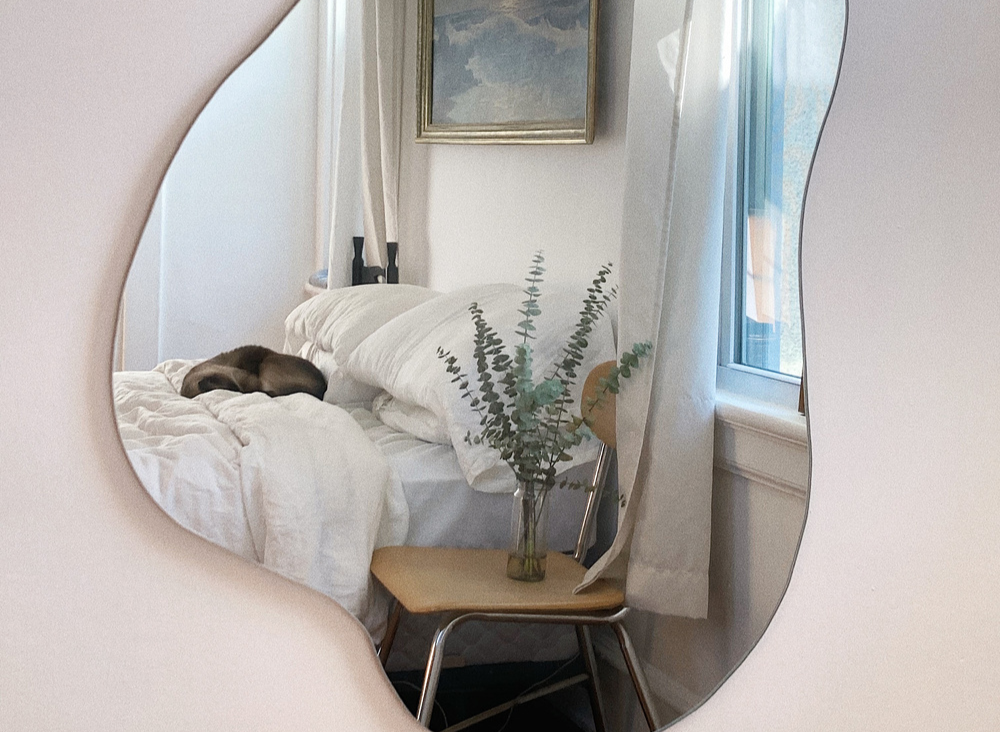 An irregular-shaped wall mirror reflects a bedroom with a cozy bed, a small chair, and a framed painting on the wall.