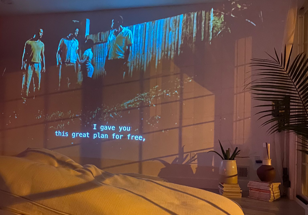 A projector displays a film onto a bedroom wall above a plush, cozy bed illuminated by warm, sunset-toned light.