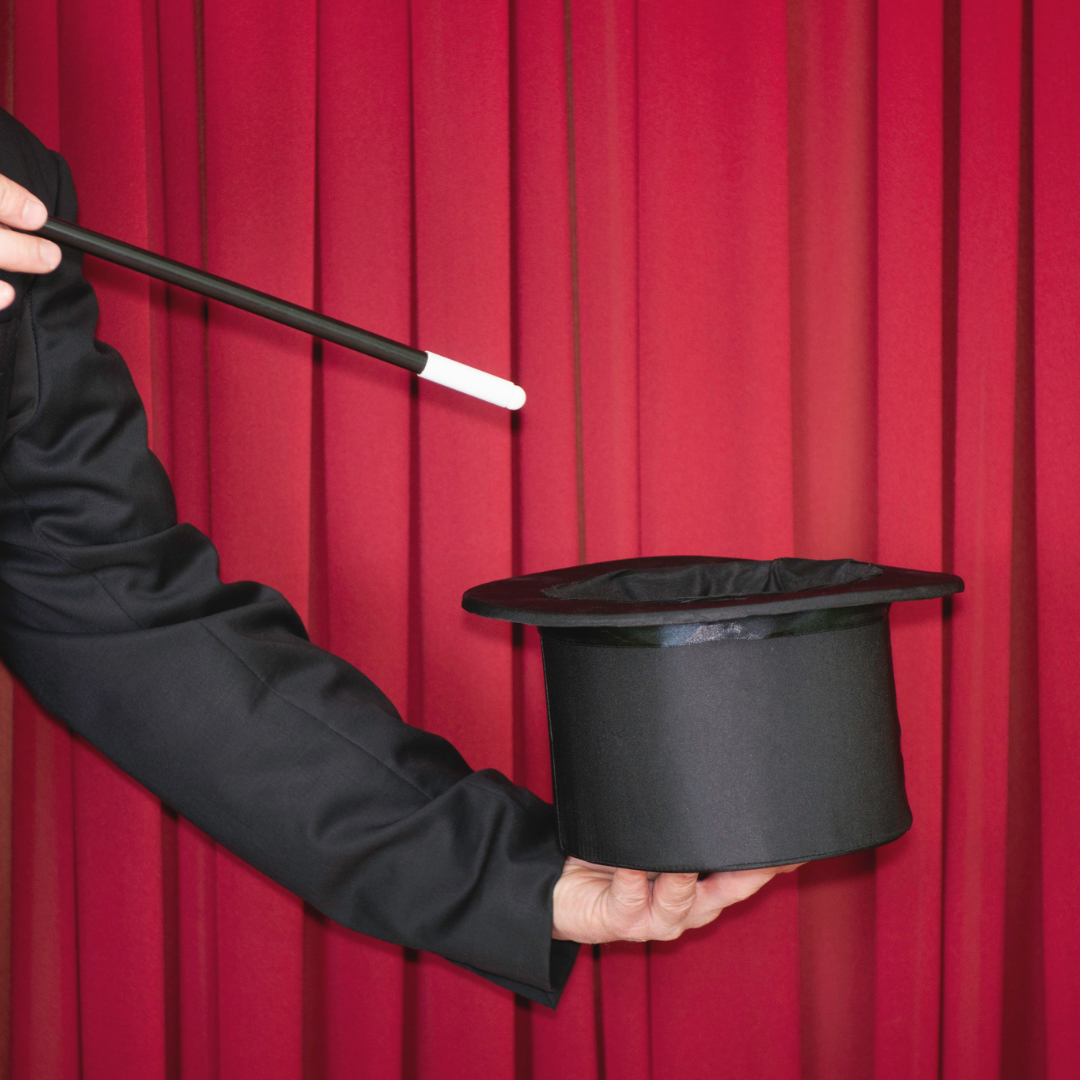 Magician holding a top hat and wand in front of a red curtain, performing a trick.