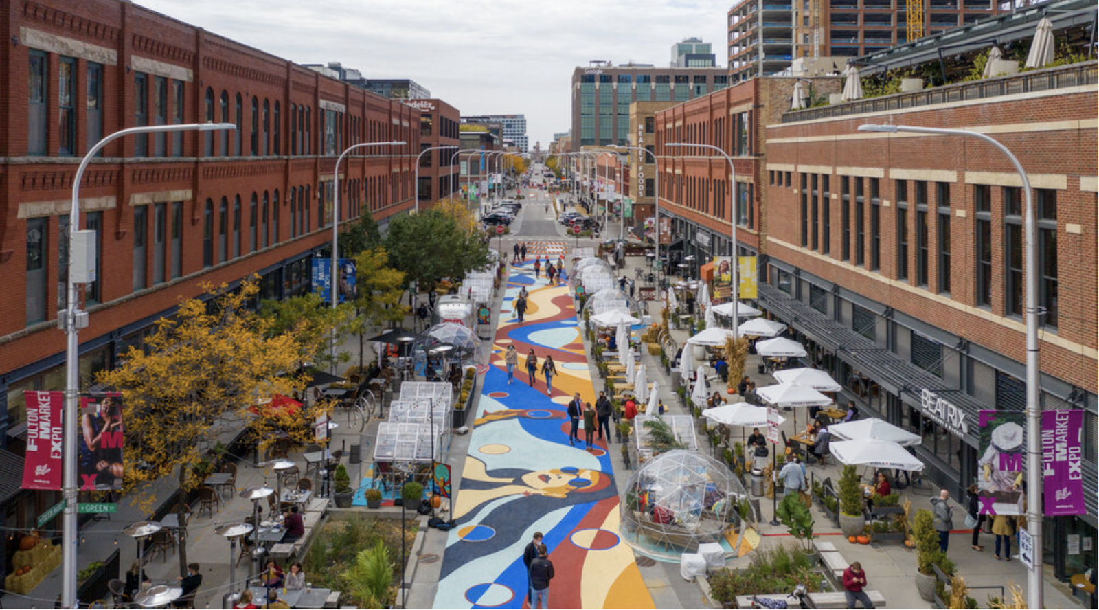 An elevated view of a pedestrian plaza with a colorful, abstract painted street mural between brick storefronts.