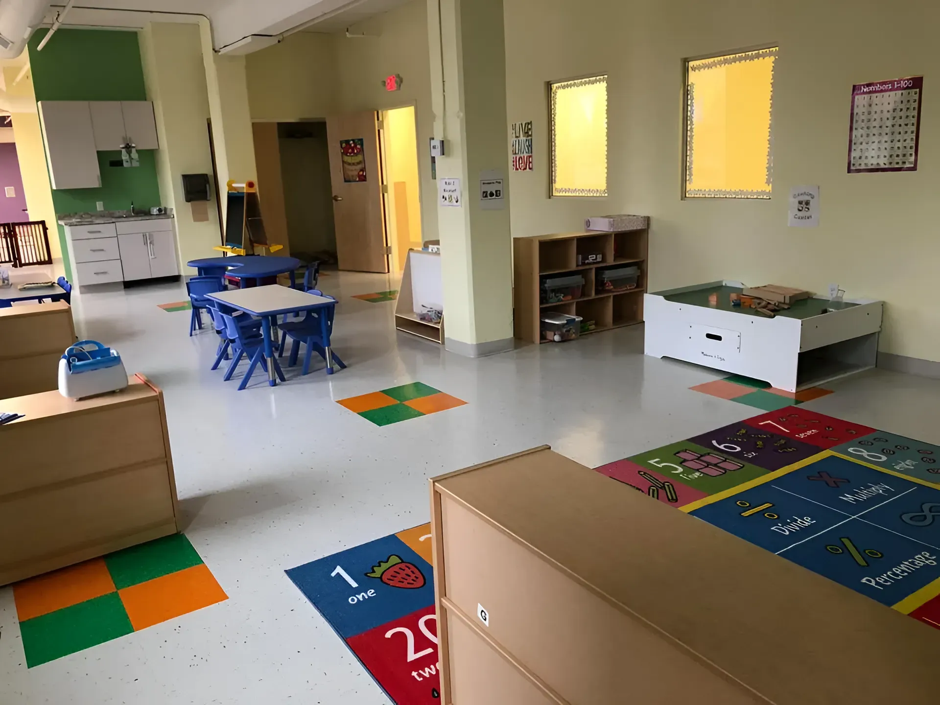 Bright classroom with colorful rugs, small tables, chairs, and a low shelf by windows.