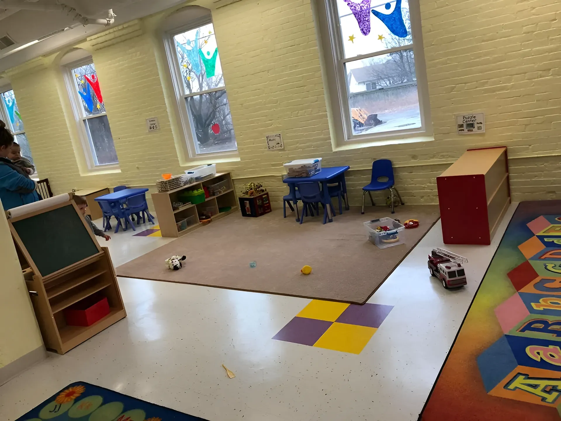 Empty preschool classroom with small tables, blue chairs, toys, and colorful floor tiles by the windows.