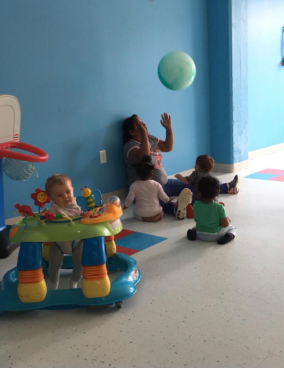 Toddlers playing in a blue room, one reaching for a floating ball near a caregiver.