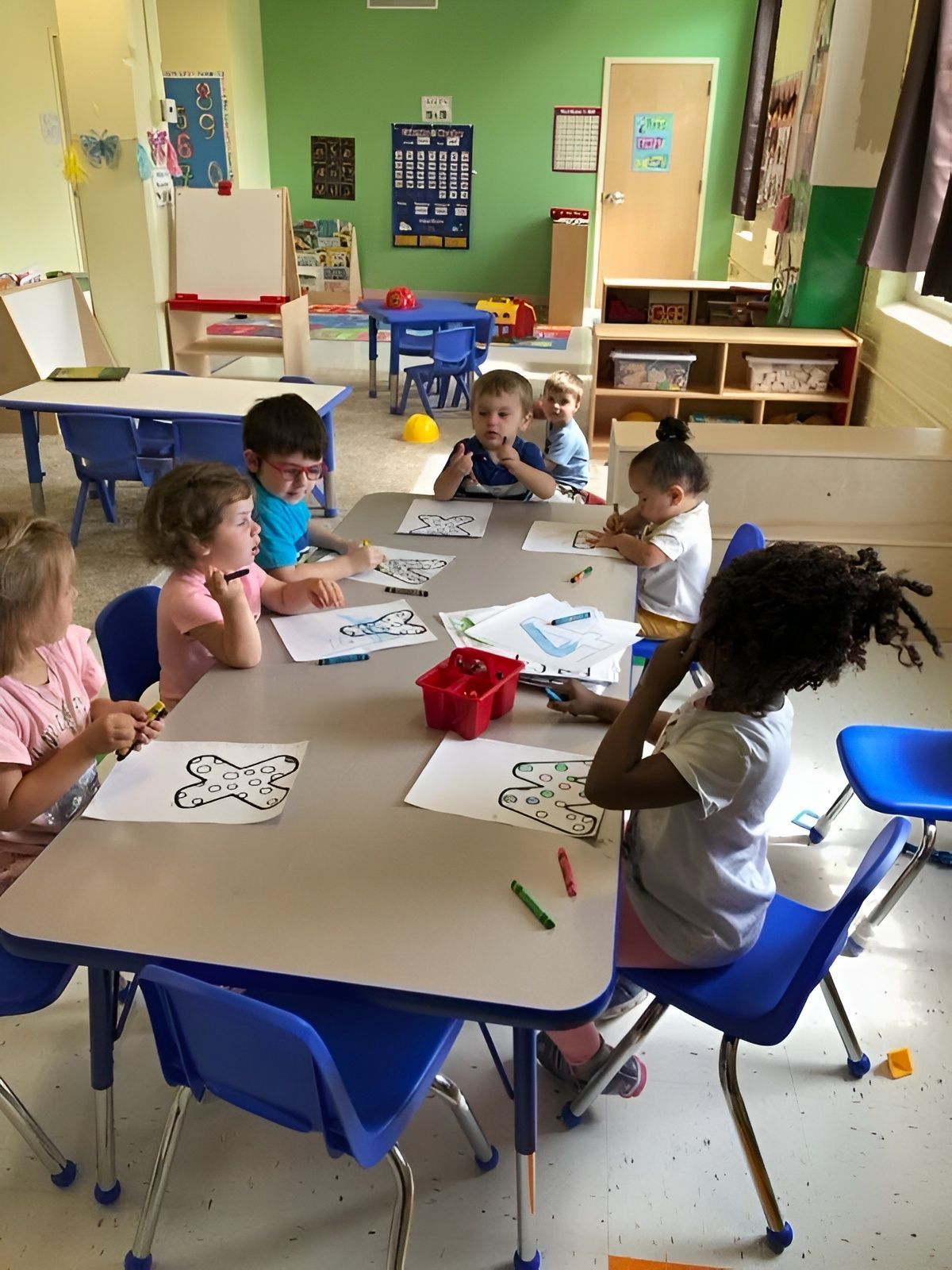Children coloring at a classroom table with paper, crayons, and markers in a bright preschool room
