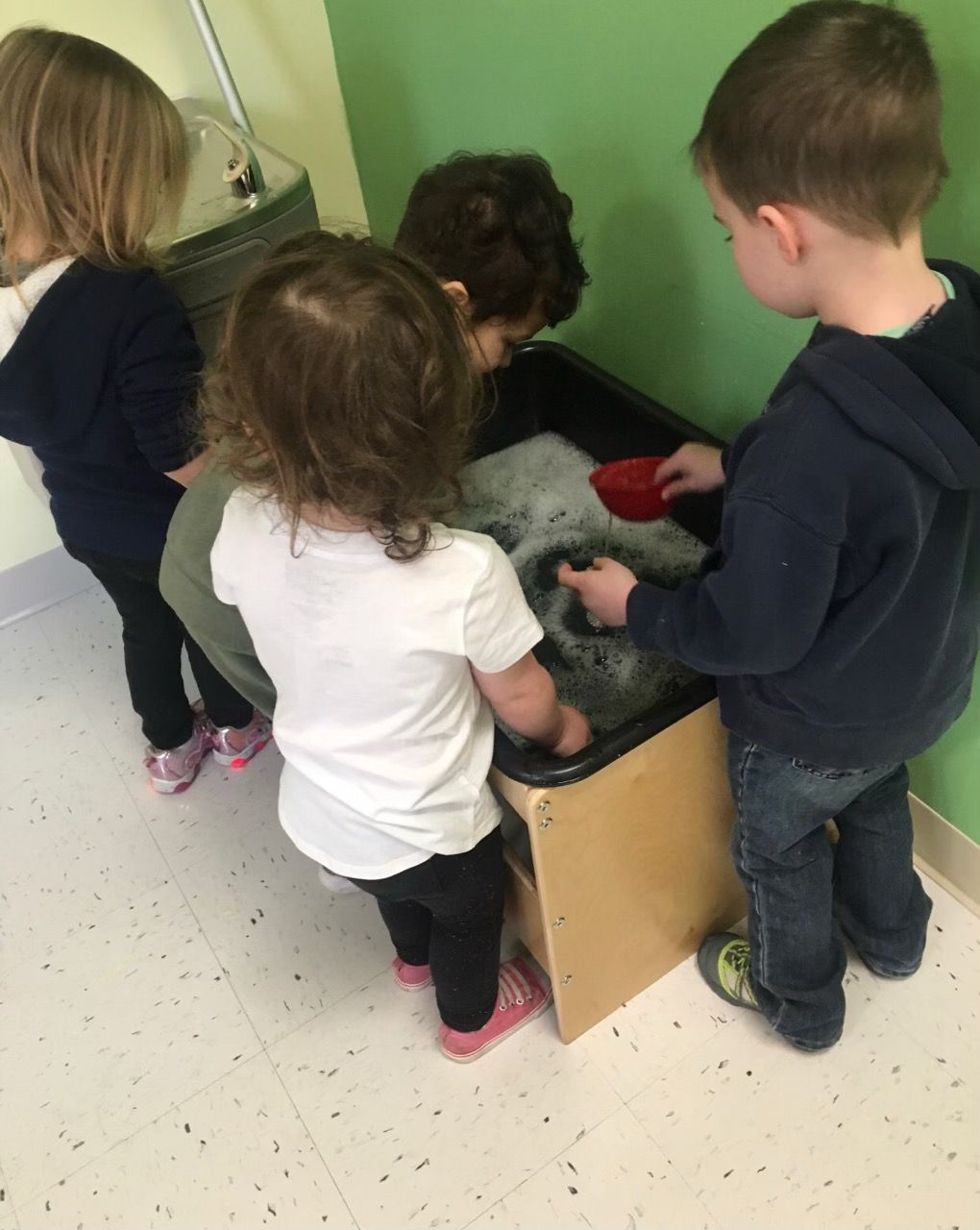 Four children stand around a sensory bin in a classroom, scooping white and red materials.