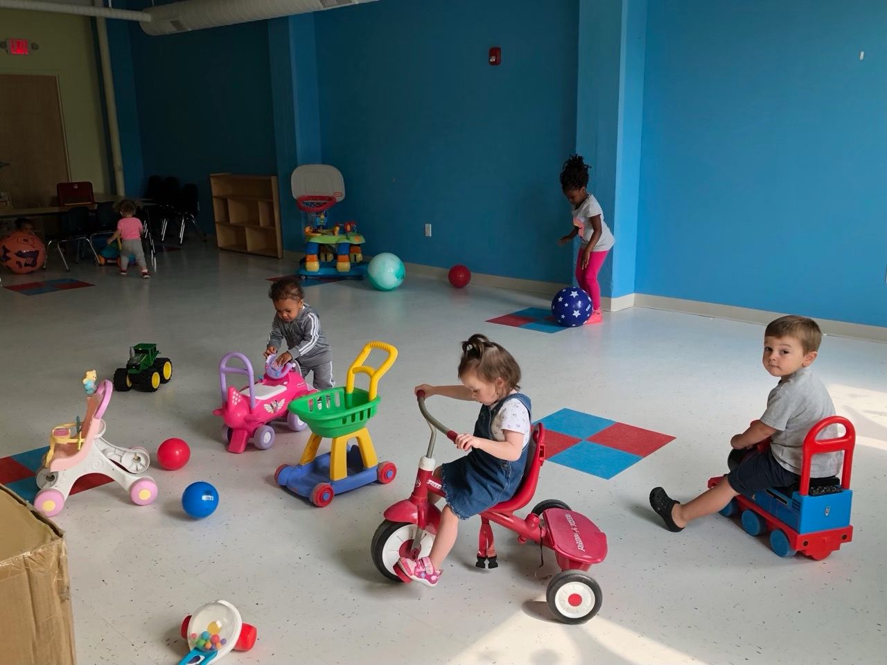 Children playing with ride-on toys in a bright indoor playroom with blue walls.