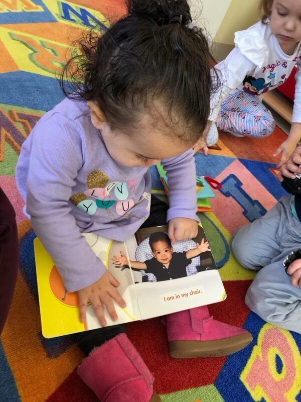Child sitting on a colorful carpet, reading a picture book with an adult nearby.