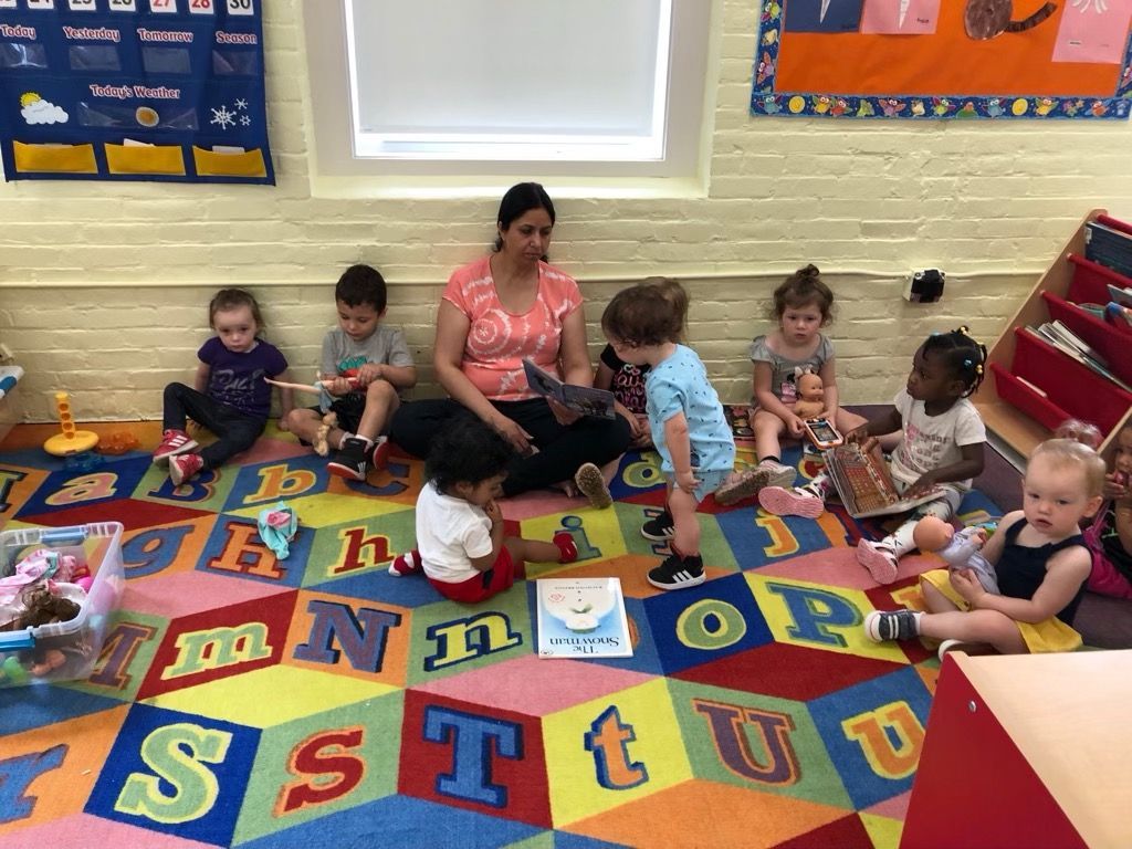 Children sitting on a colorful alphabet rug in a classroom, playing with books and toys with a teacher nearby