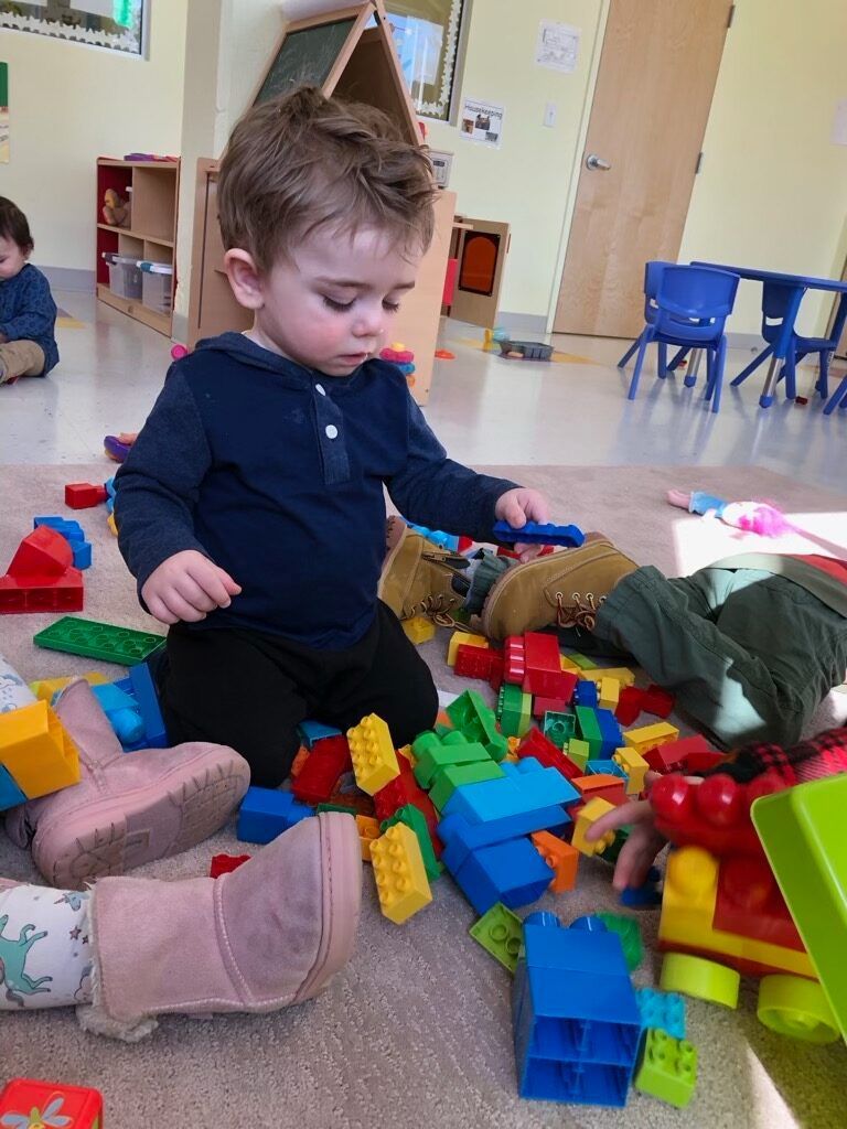 Toddler kneeling in a playroom, reaching into a pile of colorful plastic building blocks.