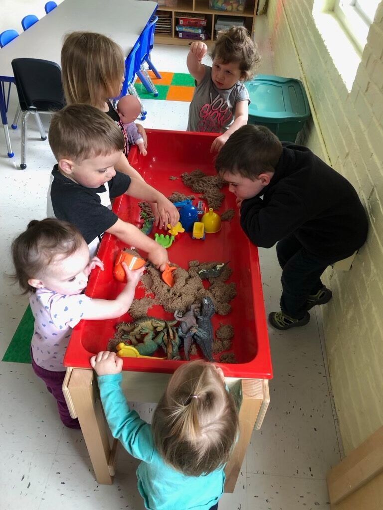Children play with toys around a red sensory table in a bright classroom.