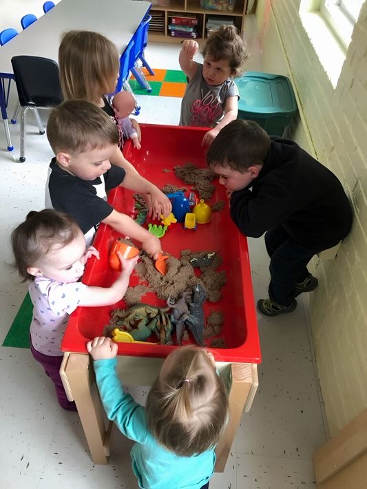 Children playing with toys around a red table in a classroom or daycare setting