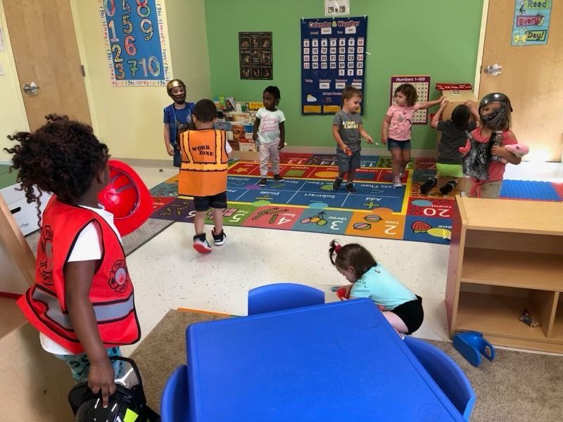 Children playing in a colorful classroom with a rug, tables, and educational wall charts.