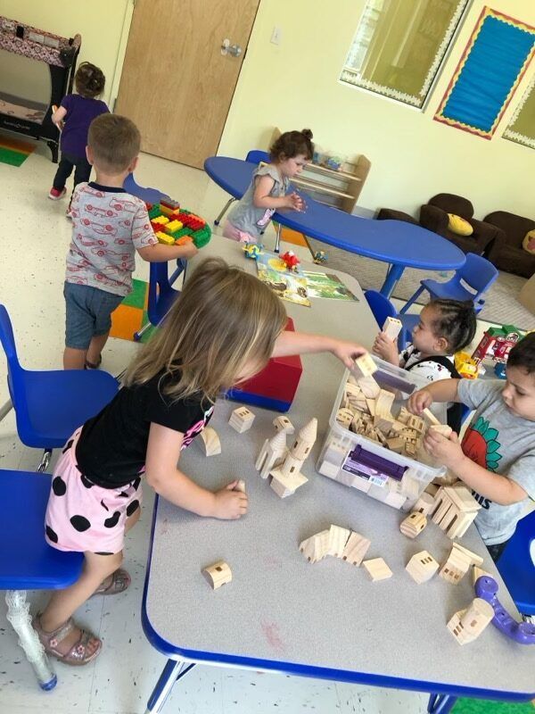 Children playing with wooden blocks at a classroom table, with colorful toys and blue chairs.