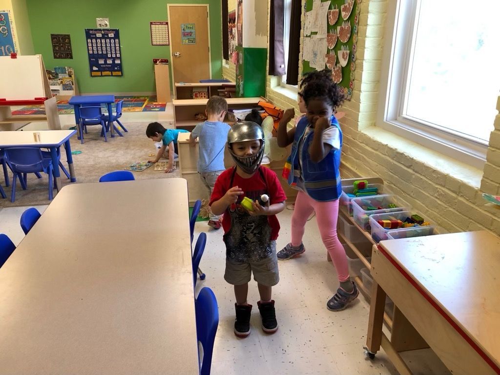Children playing in a colorful classroom with tables, chairs, and toy bins near a bright window
