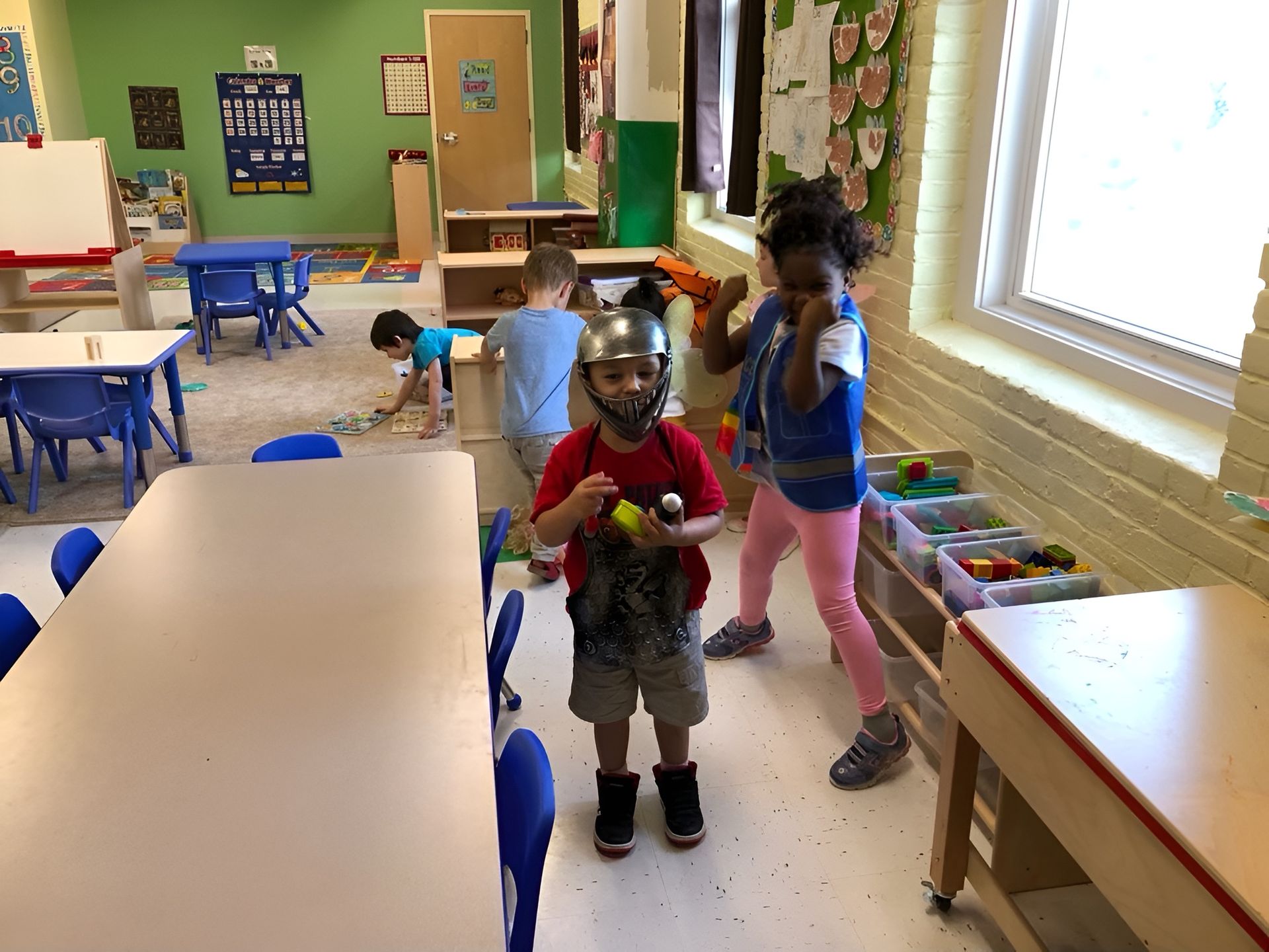 Children playing in a colorful classroom with tables, blue chairs, and shelves of toys near a sunny window.