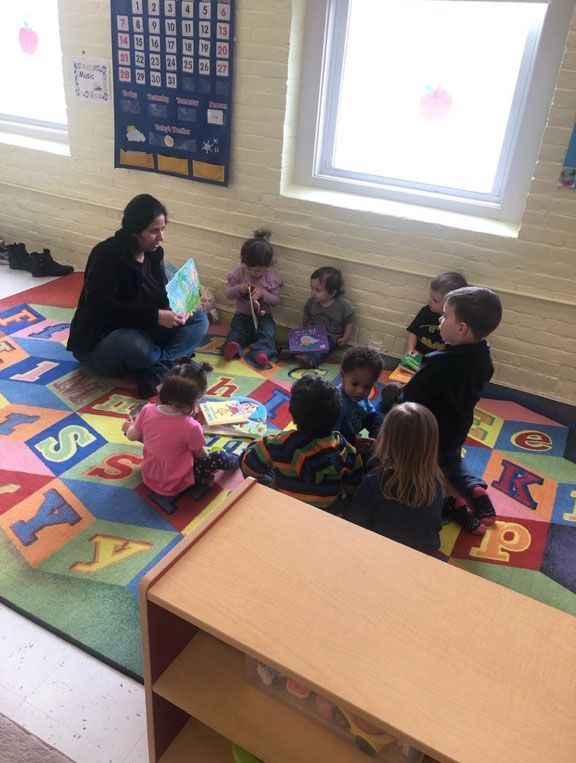 Teacher and four children sit on a colorful alphabet rug in a classroom near a bright window.
