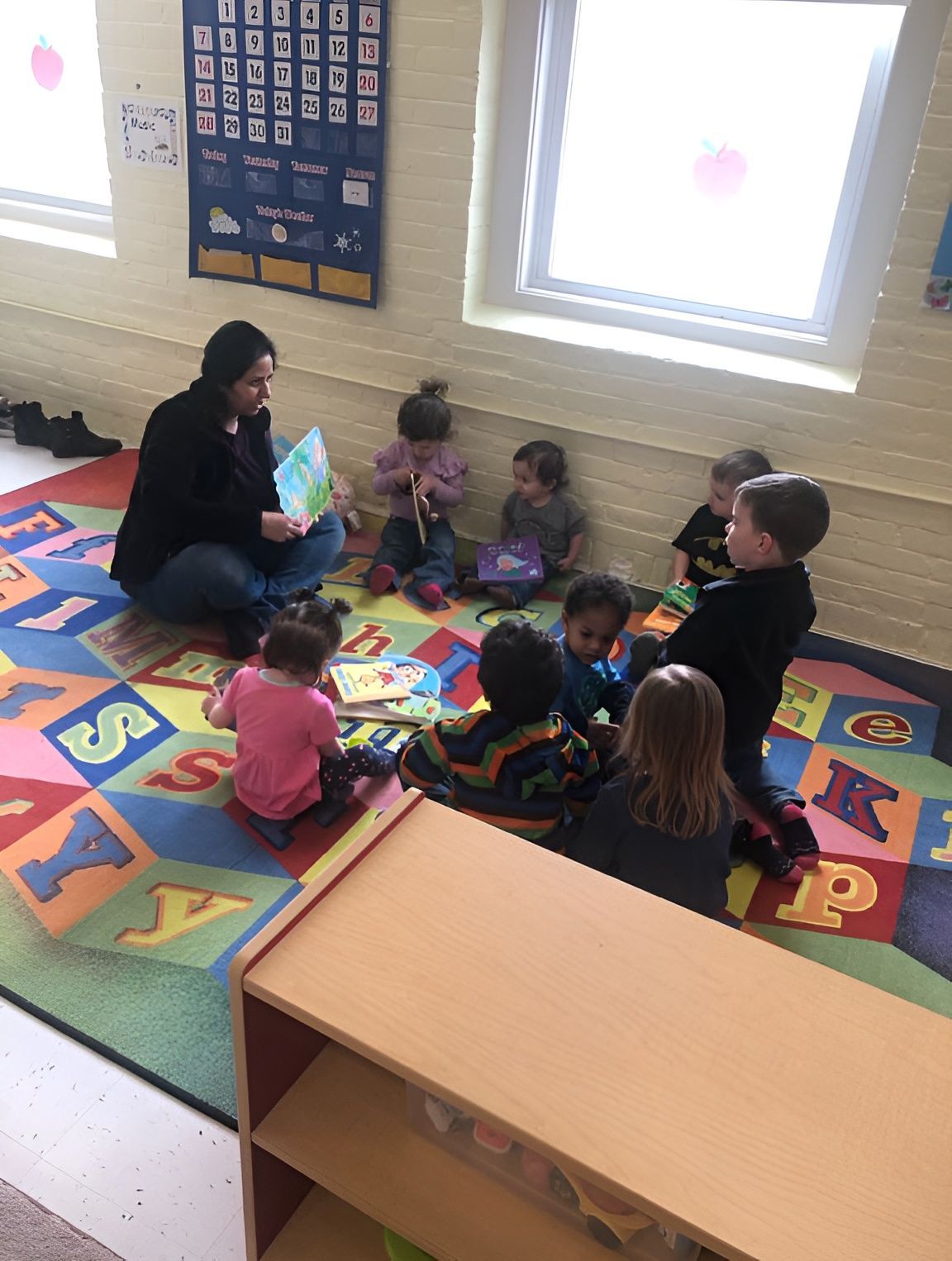 Teacher and toddlers sit on a colorful alphabet rug in a bright classroom, reading and playing together.