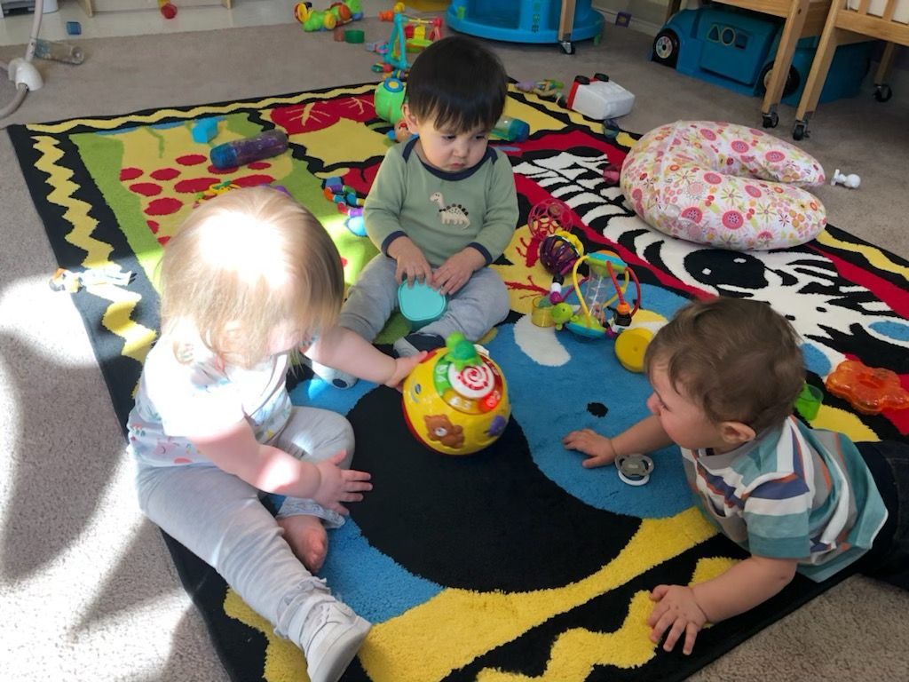 Three toddlers playing on a colorful rug with toys in a bright playroom.