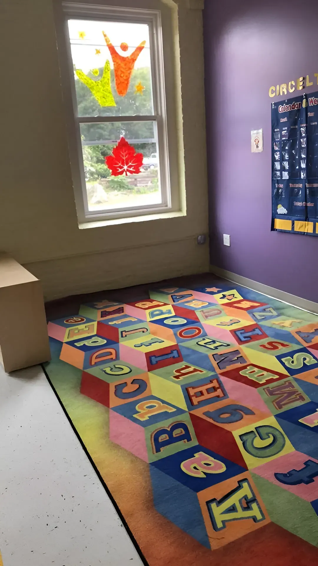 Small classroom with colorful alphabet rug, purple walls, a window, and a calendar poster.